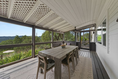 Screened covered porch with a rustic wooden dining table and chairs, lattice-beam ceiling and ceiling fan, overlooking oak trees and rolling hills