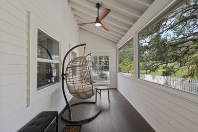 Bright screened porch with white shiplap walls and dark wood floor, two hanging wicker egg chairs beneath a modern ceiling fan, and a view of a white picket fence and sprawling oak trees.