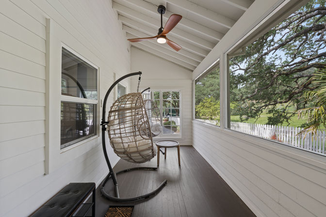 Bright screened porch with white shiplap walls and dark wood floor, two hanging wicker egg chairs beneath a modern ceiling fan, and a view of a white picket fence and sprawling oak trees.