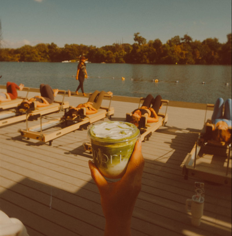 Hand holding an iced matcha latte on a sunny lakeside dock with people on reformer Pilates beds and an instructor walking by the water