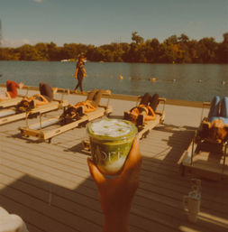 Hand holding an iced matcha latte on a sunny lakeside dock with people on reformer Pilates beds and an instructor walking by the water