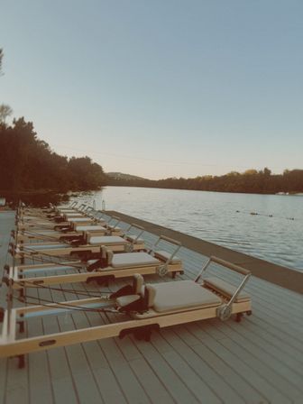 Pilates reformer machines lined up on a wooden lakeside dock at golden hour, calm water and tree-lined shoreline in the background.