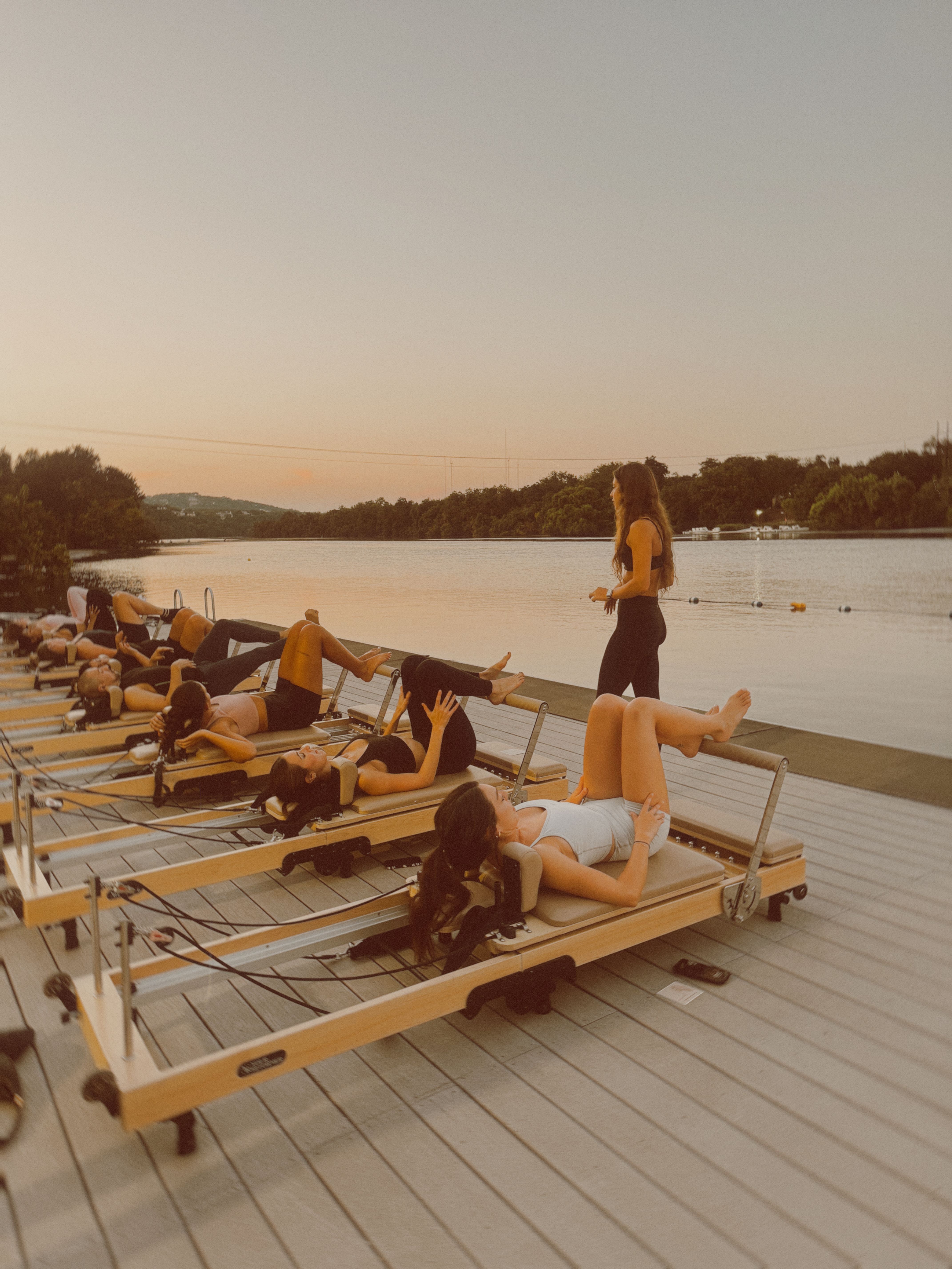 Sunset lakeside reformer Pilates class on a wooden dock — women exercising on reformer machines with an instructor standing nearby, calm water and tree-lined shore in the background.