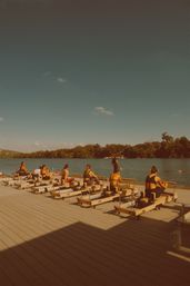 Outdoor Pilates reformer class on a lakeside wooden dock, people exercising on machines by a calm, tree-lined shore under a clear blue sky