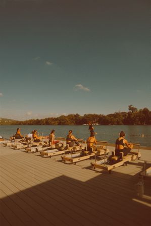 Outdoor Pilates reformer class on a lakeside wooden dock, people exercising on machines by a calm, tree-lined shore under a clear blue sky