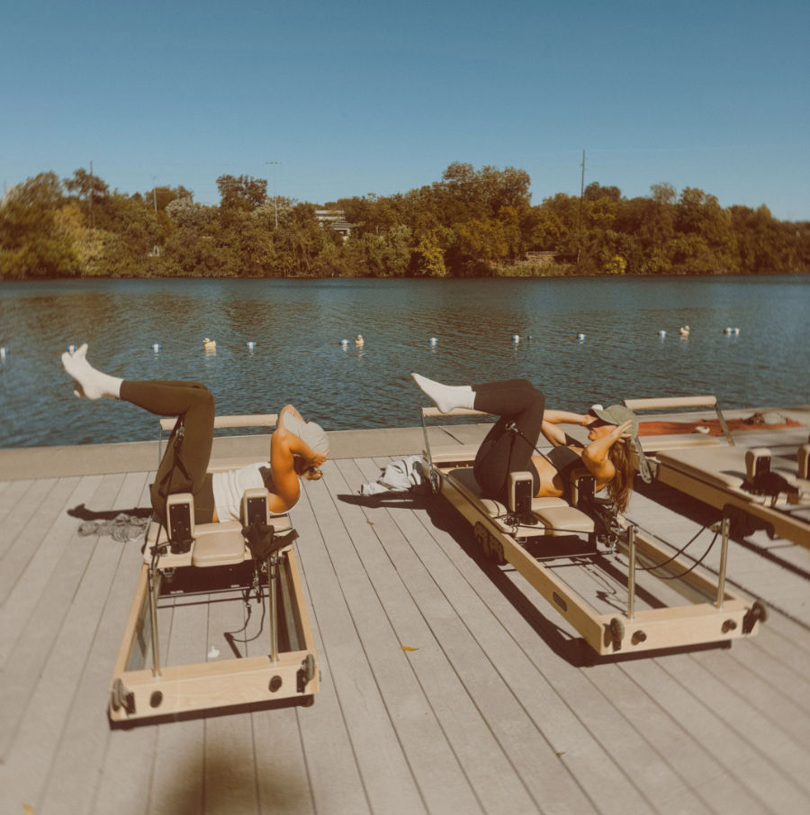 Two people doing reformer Pilates on a sunny wooden dock by a calm lake, tree-lined shoreline and clear blue sky in the background.