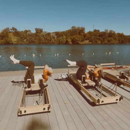 Two people doing reformer Pilates on a sunny wooden dock by a calm lake, tree-lined shoreline and clear blue sky in the background.