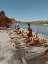 Three people doing reformer Pilates on a wooden dock by a calm lake on a sunny day, legs raised toward the sky in an outdoor lakeside fitness session
