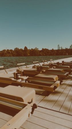 Wooden rowing machines lined up on a sunlit waterfront dock along a calm river, tree-lined opposite shore and distant urban skyline under a clear blue sky.