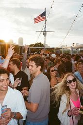 Crowded outdoor rooftop party at sunset with smiling young adults holding drinks and dancing under string lights, an American flag waving above and a city skyline in the background.
