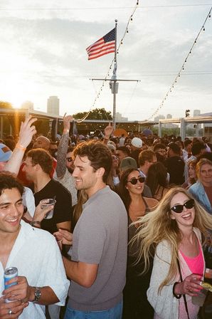 Crowded outdoor rooftop party at sunset with smiling young adults holding drinks and dancing under string lights, an American flag waving above and a city skyline in the background.