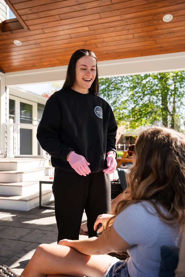 Smiling woman in a black sweatshirt and pink gloves chatting with a friend on a sunny covered backyard patio with a wooden ceiling and leafy trees.