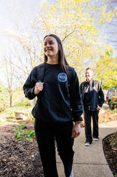 Young woman in a black sweatshirt and backpack walking down a sunny residential garden path with a smiling companion following, spring trees and blooming plants in the background.