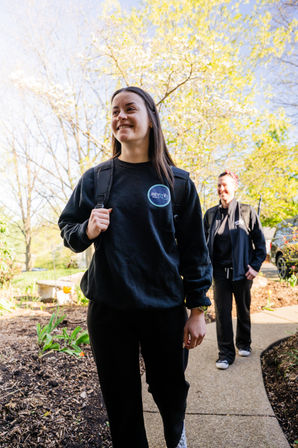 Two smiling women walking along a sunny suburban sidewalk in spring, wearing black casual outfits and backpacks amid blooming trees and tulips.