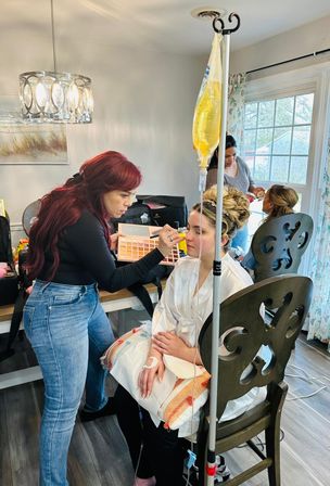 Makeup artist applying glam makeup to a seated bride in a white robe during an at-home getting-ready session, IV drip bag on a pole nearby and another stylist working in the background.