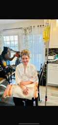 Smiling woman in a white satin robe with an updo and headband sits in a bright home kitchen with floral curtains, getting hair and makeup while a stylist works on another client; an IV drip bag on a stand is visible beside her.