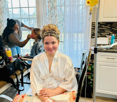 Bridal getting-ready scene in a sunlit kitchen: smiling woman in a satin robe and elegant updo sits with an IV drip and a cosmetic pad on her hand while a makeup artist touches up another woman by a window with floral curtains and styling tools on the counter.