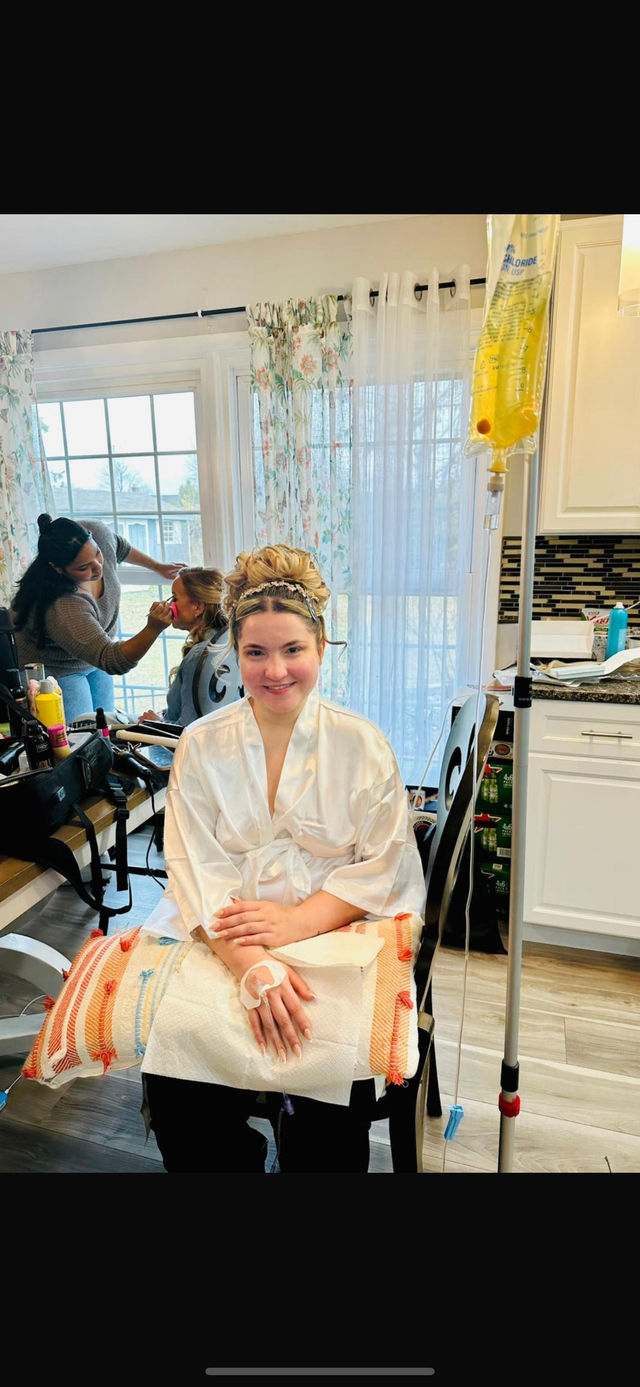 Bridal getting-ready scene in a sunlit kitchen: smiling woman in a satin robe and elegant updo sits with an IV drip and a cosmetic pad on her hand while a makeup artist touches up another woman by a window with floral curtains and styling tools on the counter.