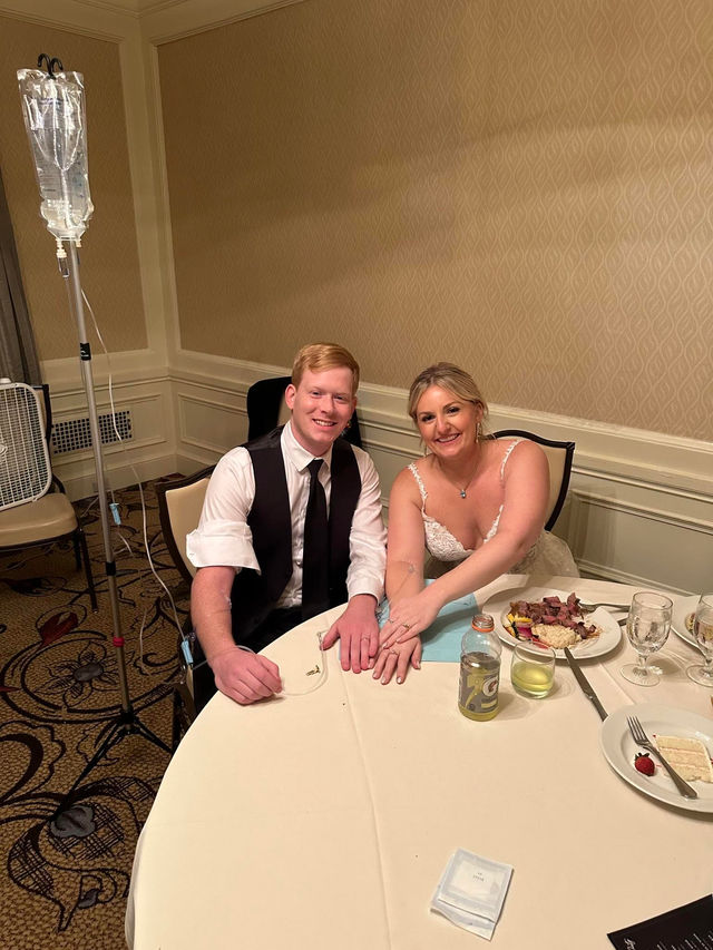 Smiling bride in a lace gown and groom in a black vest and tie sit at a round hotel ballroom wedding reception table with plates and drinks, an IV pole and fluid bag standing beside them.