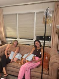 Two smiling women receiving at-home IV hydration drips while relaxing on a blush tufted sofa in a cozy living room, IV pole and clear infusion bags visible