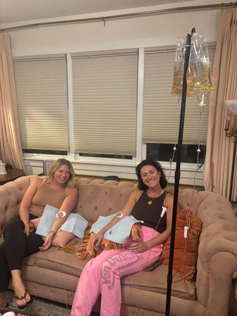 Two smiling women receiving at-home IV hydration drips while relaxing on a blush tufted sofa in a cozy living room, IV pole and clear infusion bags visible
