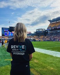 Person with shoulder-length blonde hair wearing a black T-shirt with IV therapy branding and 'Pittsburgh' on the back stands on the football sideline watching a packed Pittsburgh stadium, scoreboard and partly cloudy sky.