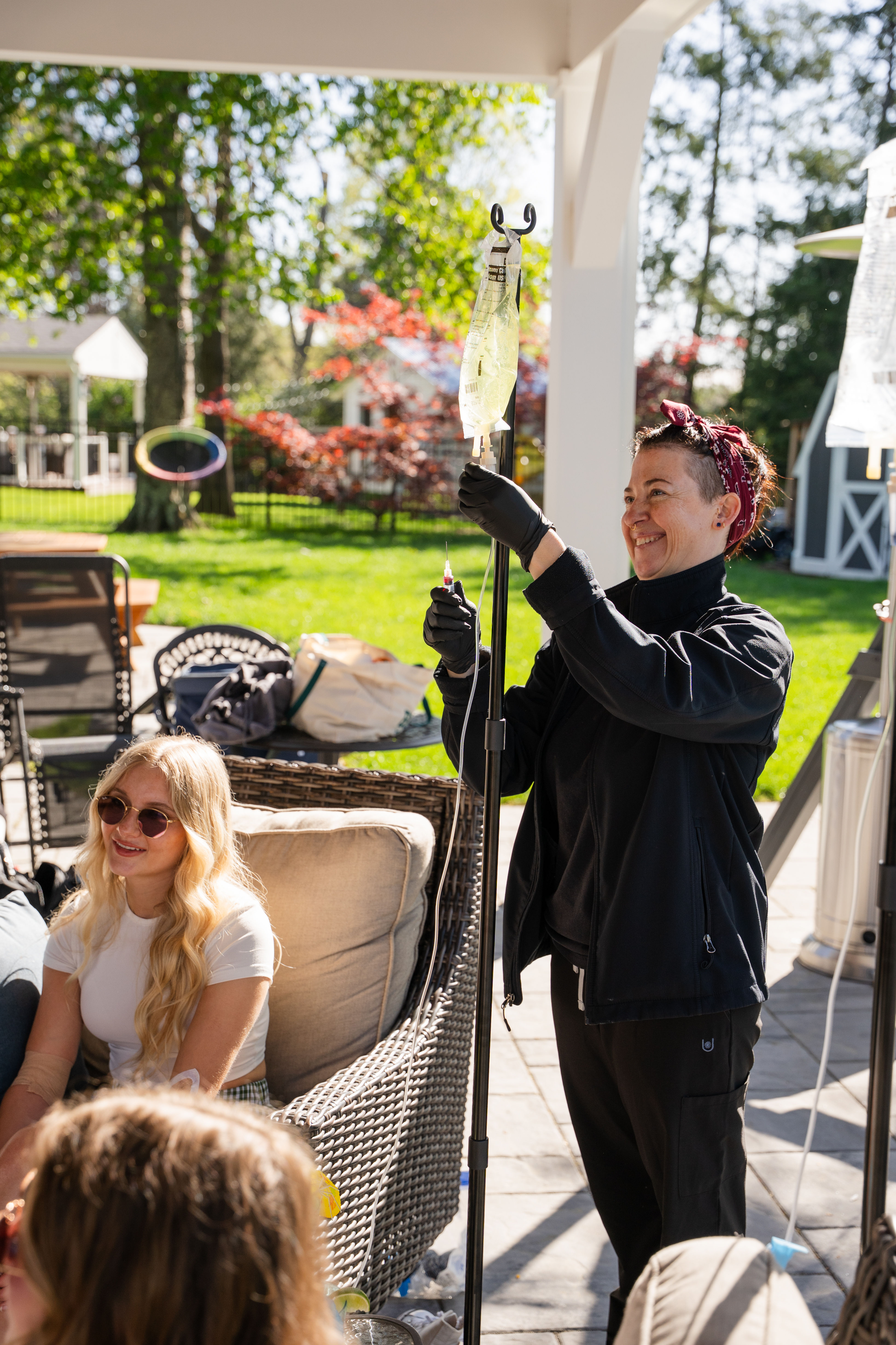 Smiling caregiver adjusts an IV drip on a stand while friends relax on wicker patio furniture in a sunny suburban backyard with green lawn and colorful trees.