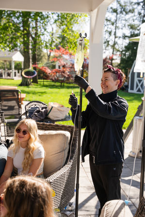 Smiling caregiver adjusts an IV drip on a stand while friends relax on wicker patio furniture in a sunny suburban backyard with green lawn and colorful trees.