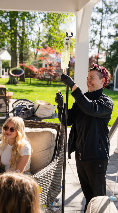 Smiling caregiver adjusts an IV drip on a stand while friends relax on wicker patio furniture in a sunny suburban backyard with green lawn and colorful trees.