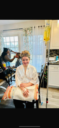Smiling bride in a white satin robe sits in a bright kitchen during wedding morning preparations — hairstylist applying makeup in the background and an IV hydration bag on a stand beside her.