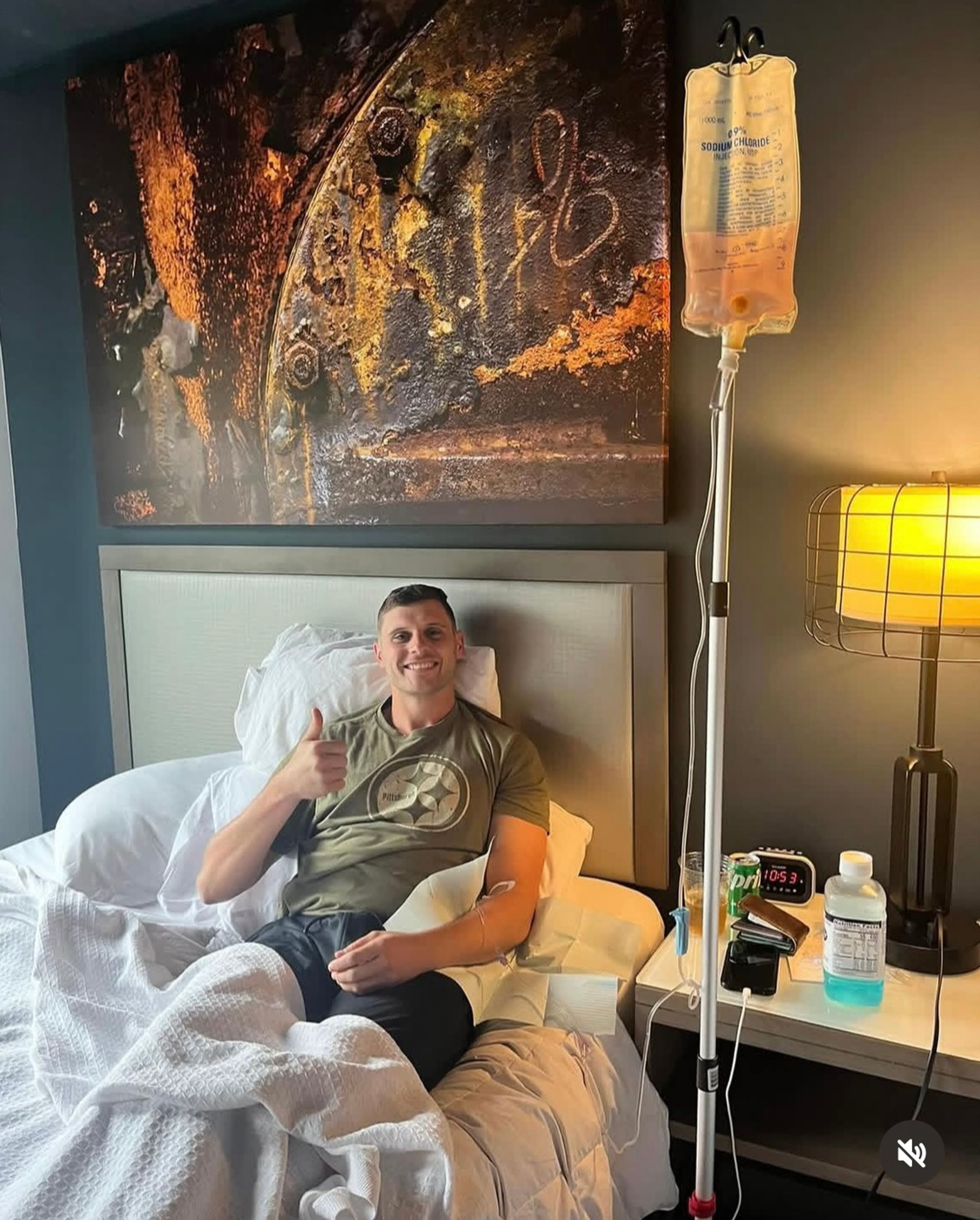 Smiling man reclining in a hotel bed with an IV drip in his arm, giving a thumbs-up; bedside lamp, alarm clock and water bottle on the nightstand, abstract wall art above.