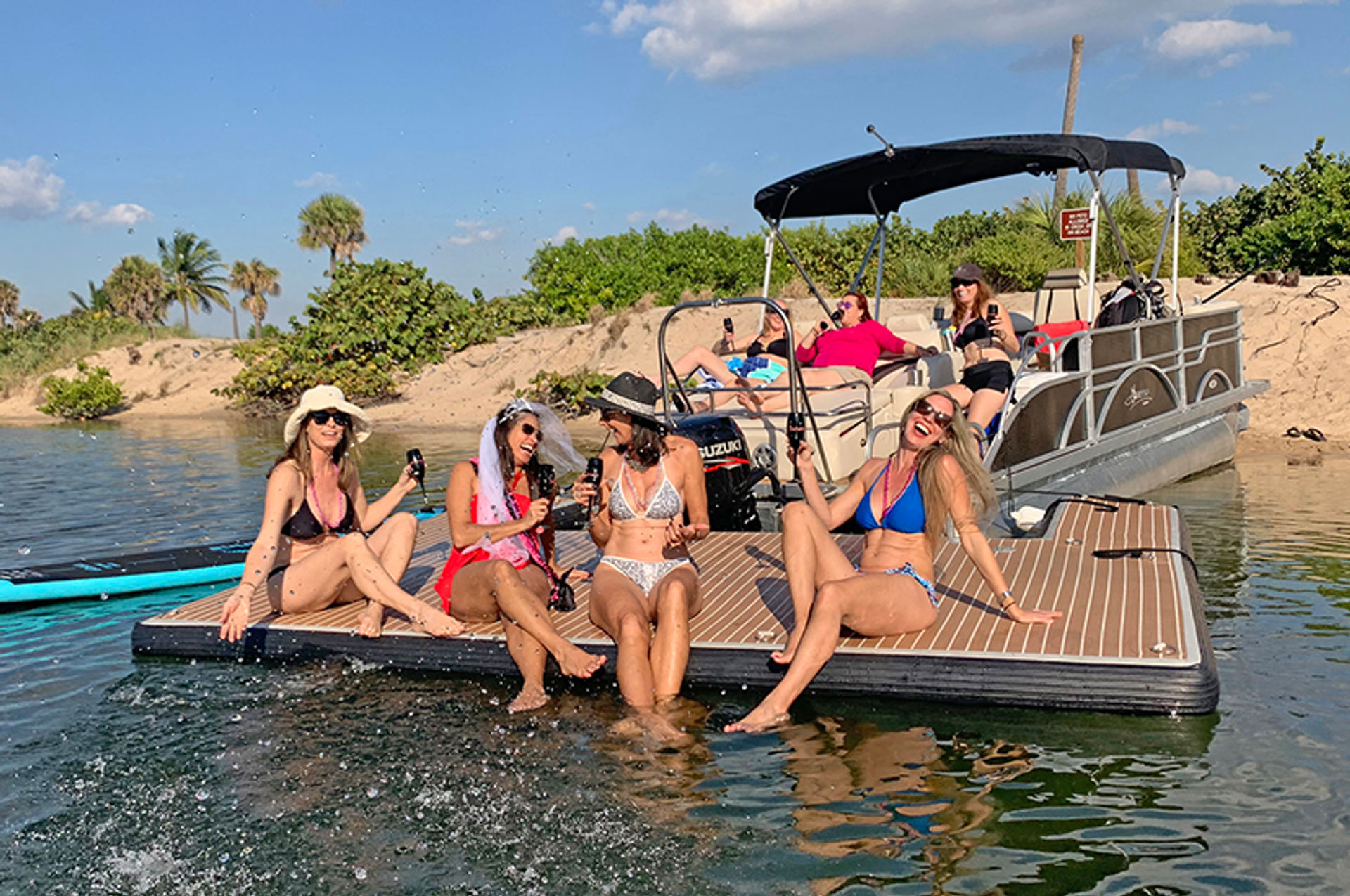 Friends in swimsuits laughing and toasting drinks on a pontoon boat platform near a sandy, palm-lined beach — sunny bachelorette celebration on the water.