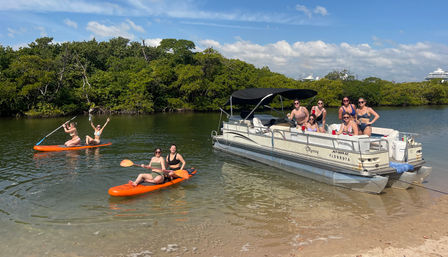 Group enjoying a sunny day on a pontoon boat and bright orange kayaks in a mangrove-lined coastal inlet by a sandy shore.