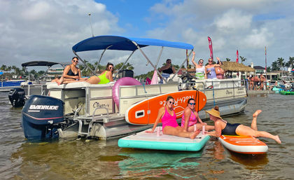 Friends enjoying a sunny beach party on a pontoon boat and paddleboards in a palm-lined coastal bay with colorful inflatables.