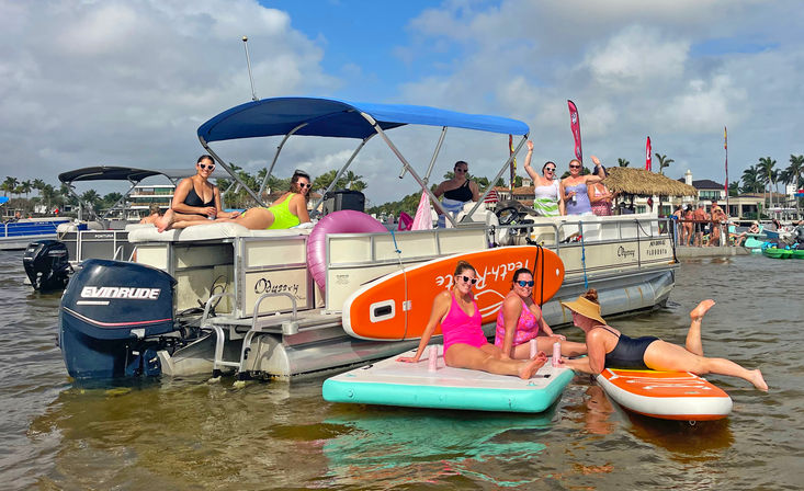 Friends enjoying a sunny beach party on a pontoon boat and paddleboards in a palm-lined coastal bay with colorful inflatables.