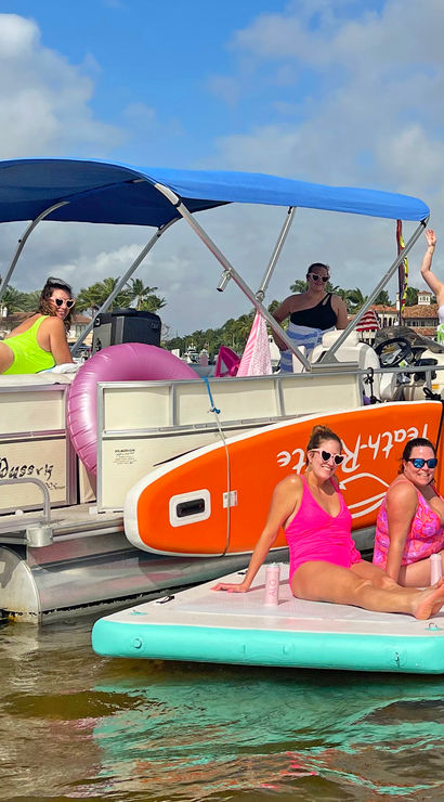 Friends enjoying a sunny beach party on a pontoon boat and paddleboards in a palm-lined coastal bay with colorful inflatables.