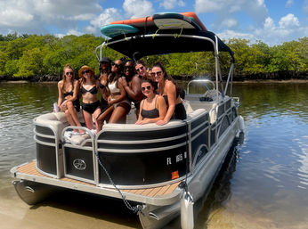 Smiling group of women in swimsuits posing on the bow of a pontoon boat in shallow water by mangroves on a sunny Florida waterway, paddleboards secured on the boat canopy.