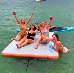 Group of friends in bikinis cheering with drinks on an inflatable floating mat near a paddleboard in clear green coastal water