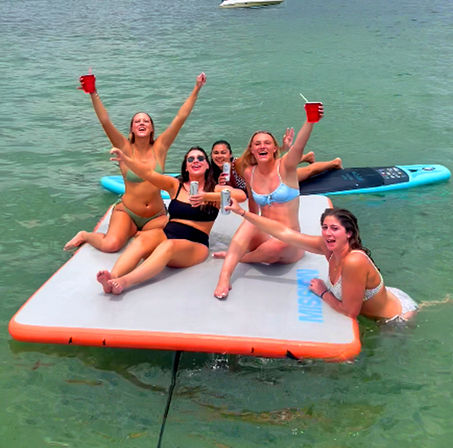 Group of friends in bikinis cheering with drinks on an inflatable floating mat near a paddleboard in clear green coastal water