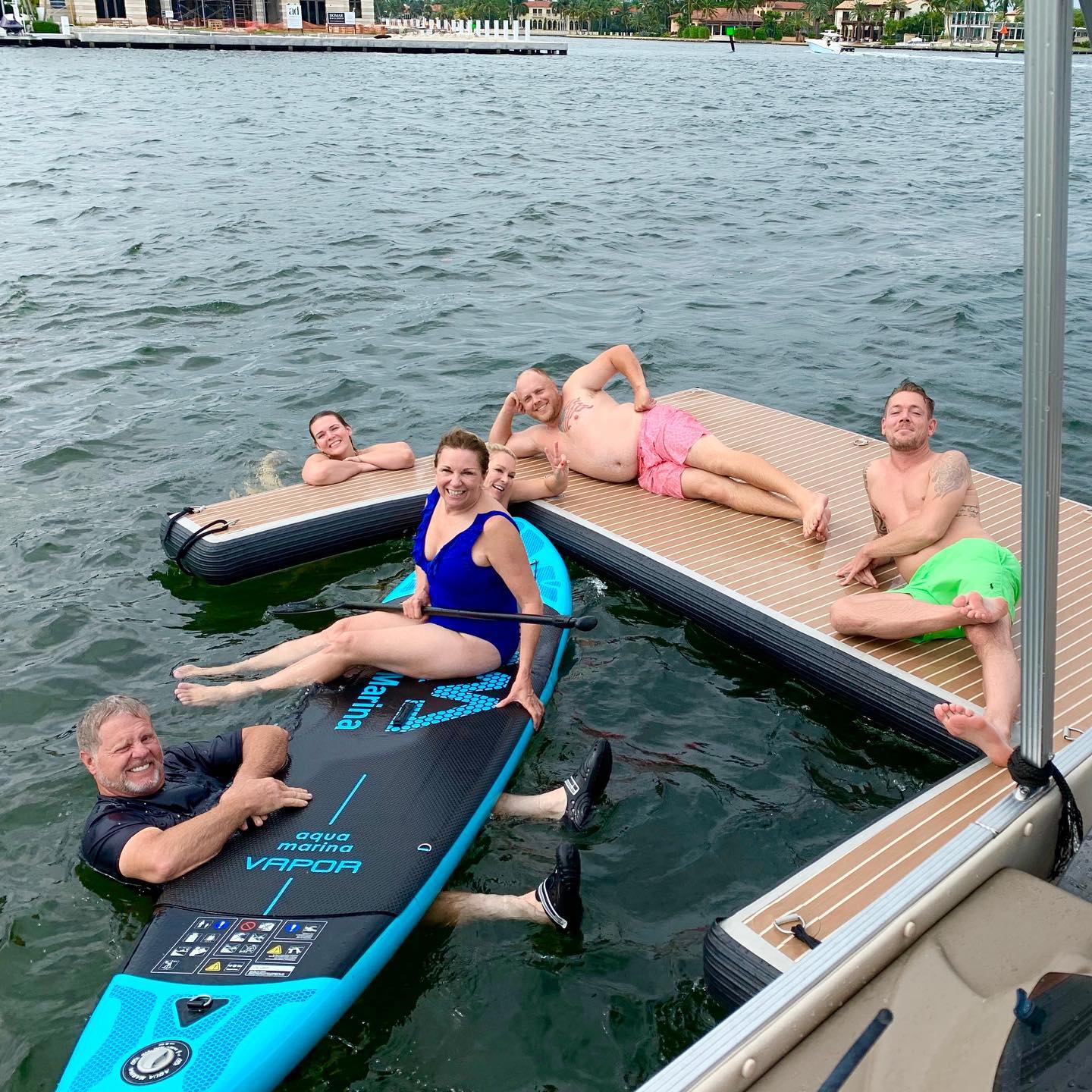 Smiling adults lounging on a stand-up paddleboard and floating dock at a waterfront marina — colorful swimsuits and summer boating fun.
