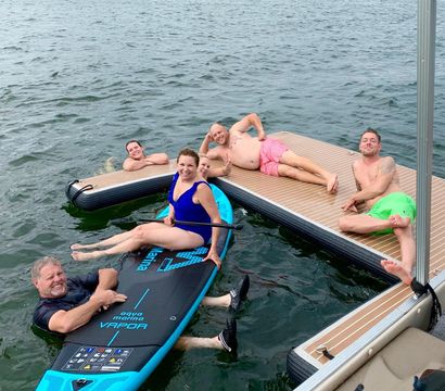 Smiling adults lounging on a stand-up paddleboard and floating dock at a waterfront marina — colorful swimsuits and summer boating fun.