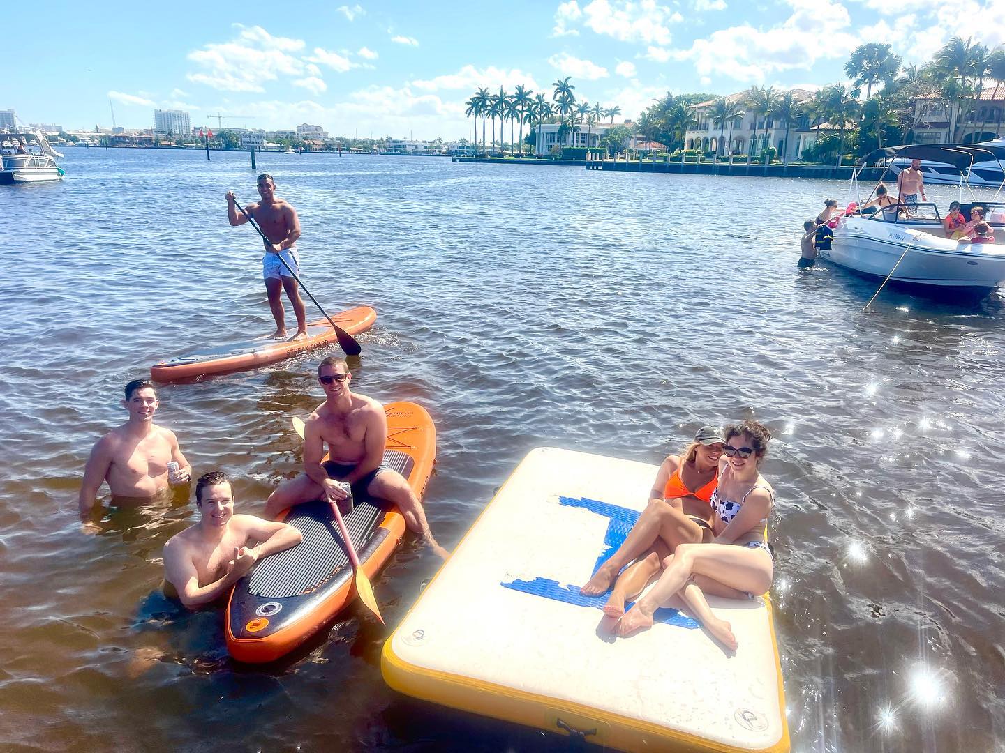Smiling friends paddleboarding and lounging on an inflatable water platform by palm-lined waterfront mansions and boats on a sunny coastal Florida waterway