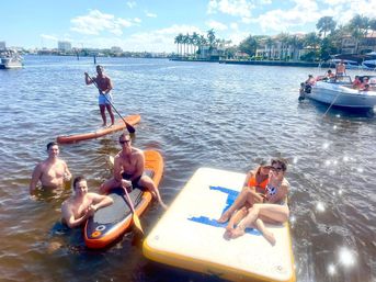 Smiling friends paddleboarding and lounging on an inflatable water platform by palm-lined waterfront mansions and boats on a sunny coastal Florida waterway