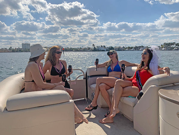 Four women laughing on a pontoon boat near a palm-lined waterfront, wearing swimsuits and holding drinks; bachelorette in veil and sash under a sunny, cloud-dotted sky.