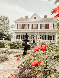 Charming white two-story home with a wrap porch and brown shutters, brick walkway and red roses in the front yard leading to a black tiered fountain