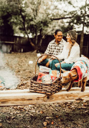 Backyard autumn picnic scene: wicker basket of s'mores and snacks on a wooden beam, a small goat in a colorful sweater sniffing the basket, and a smiling couple blurred by a campfire in the background.