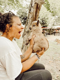 Laughing woman holding a fluffy brown baby goat (kid) with pale blue eyes in a shaded outdoor farmyard petting area with trees, wooden fence and picnic tables in the background.