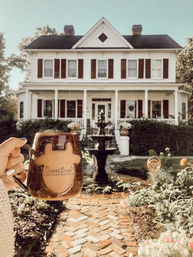Cozy morning scene: hand holding a brown ceramic coffee mug in front of a white Victorian farmhouse with red shutters, wraparound porch, brick walkway and a tiered garden fountain in the sunny front yard.