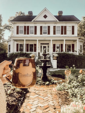 Cozy morning scene: hand holding a brown ceramic coffee mug in front of a white Victorian farmhouse with red shutters, wraparound porch, brick walkway and a tiered garden fountain in the sunny front yard.