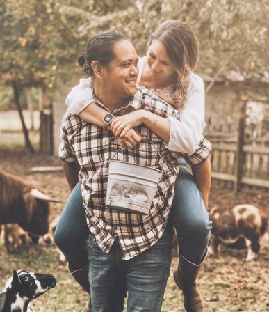Smiling couple on a rustic country farm: man in plaid gives woman a piggyback as she holds a metal bucket, goats and cows grazing nearby.